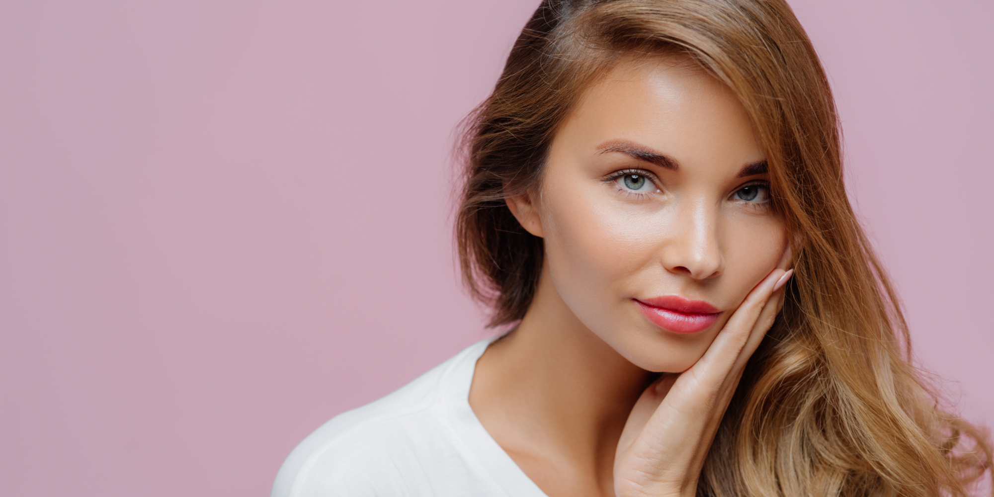 Woman with a soft smile and hand on her cheek against a pink background